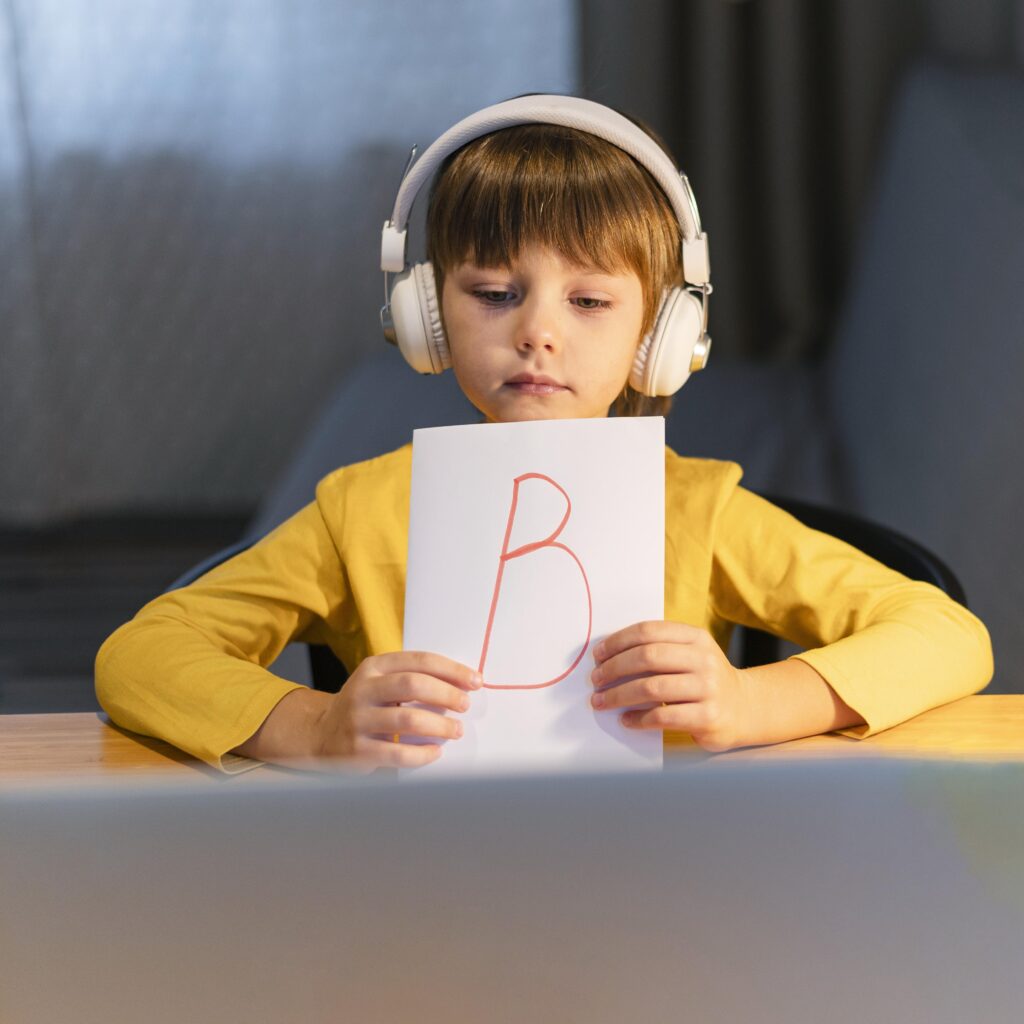 Child in yellow shirt with headphones holding up a paper with a red letter "B" drawn on it.